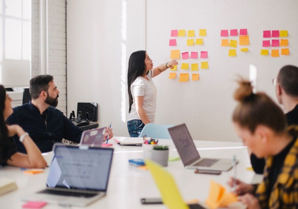 Team brainstorming session with sticky notes on wall, woman presenting ideas to colleagues, laptops on table, collaborative environment focused on brand management strategies.